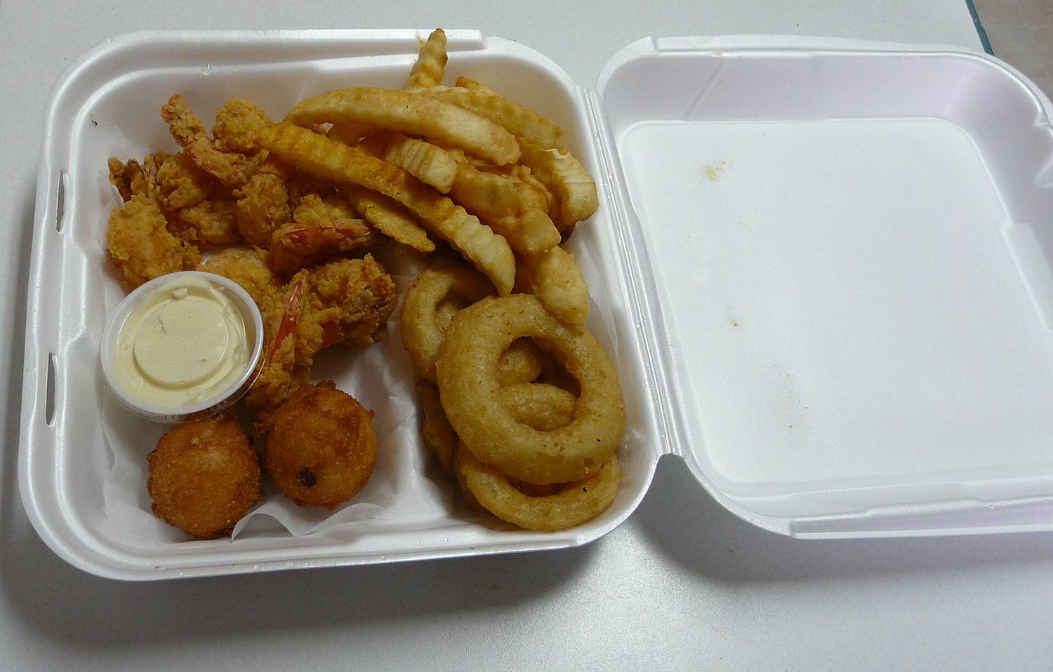 A takeout box with fried shrimp, onion rings and hush puppies